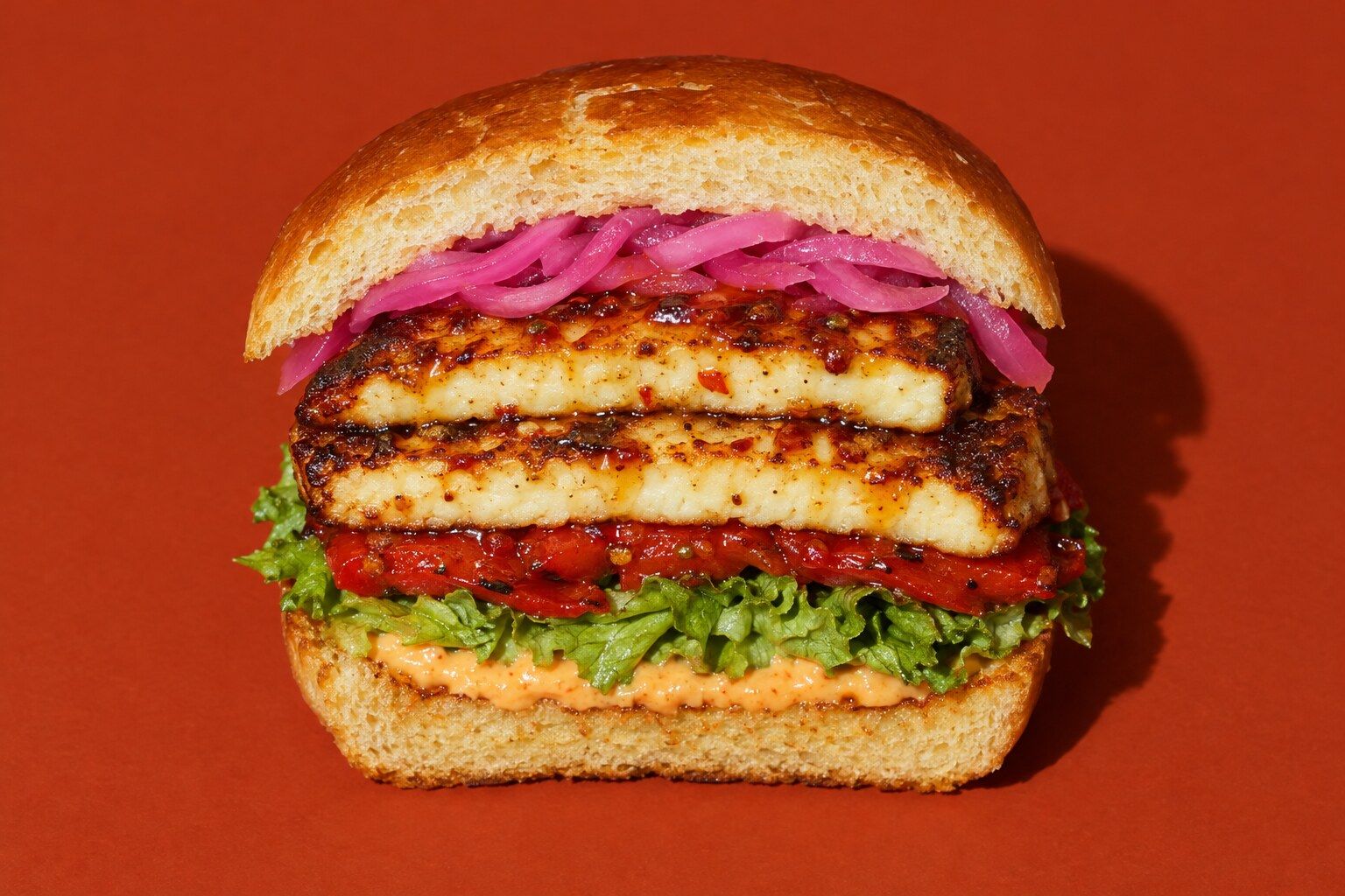 En hög burgare i briochebröd med två glaserade skivor stekt grillost, rosa picklad rödlök, grillad paprika, sallad och orange majonnäs.